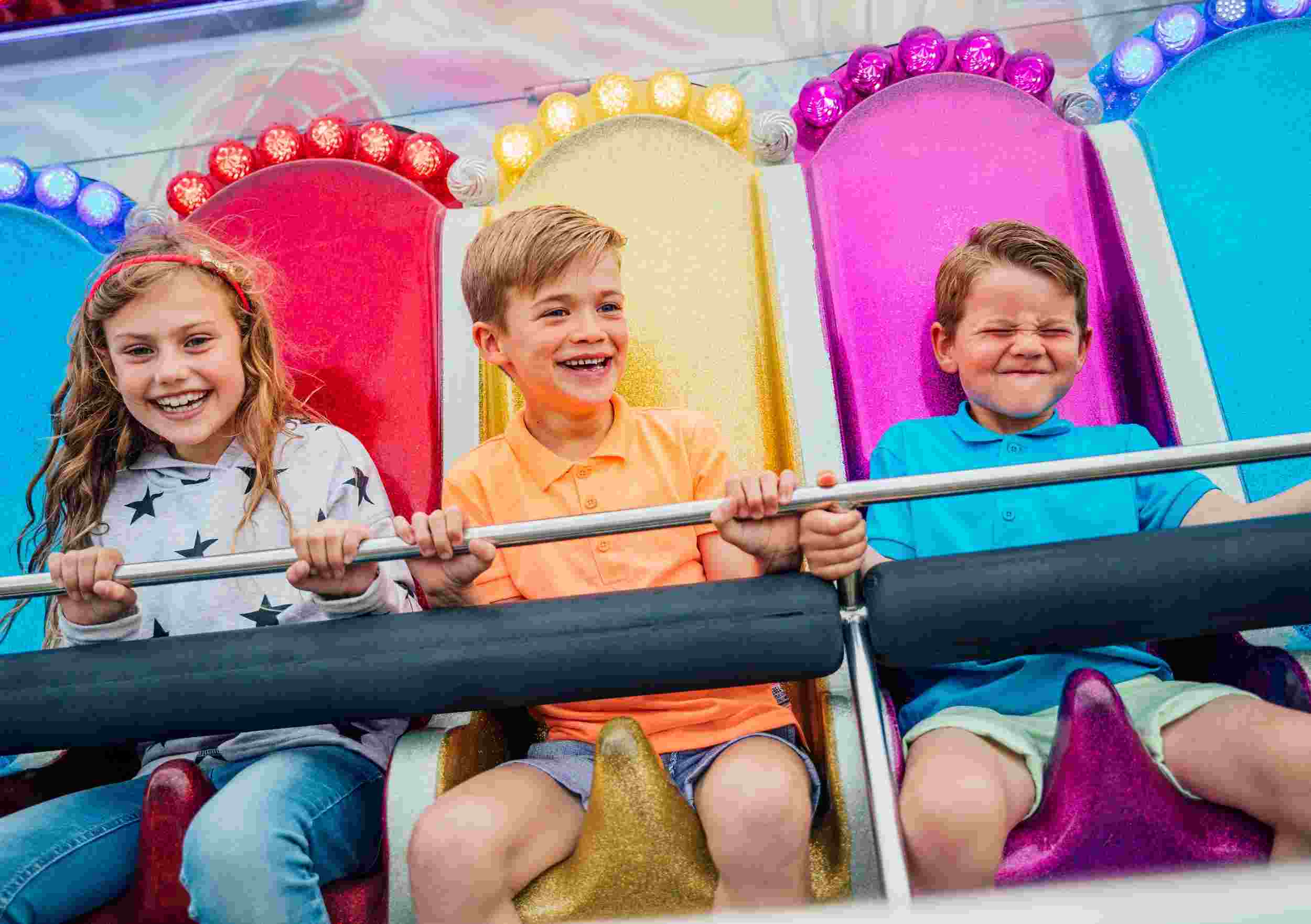 Three children smiling and laughing on a colorful fairground ride, engaged in summer activities